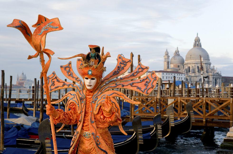 Male mask in creative orange costume at carnival in Venice, Italy. Landing stage with gondolas and Santa Maria della Salute cathedral in the background.