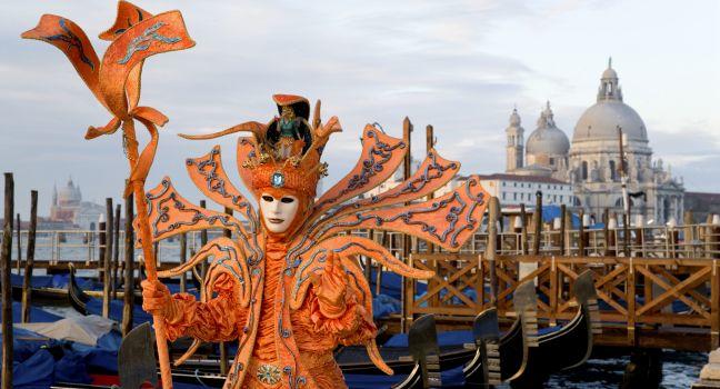 Male mask in creative orange costume at carnival in Venice, Italy. Landing stage with gondolas and Santa Maria della Salute cathedral in the background.